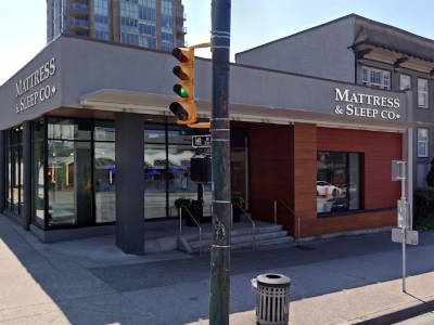 A storefront for Mattress & Sleep Co. on a South Granville Vancouver city street, with large glass windows, a gray and wood facade, a traffic light, sidewalk, and nearby buildings.
